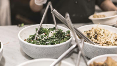Image of table set with serving bowls, one with kale another with cauliflower, and tongs for serving and an out-of-focus person at end of table